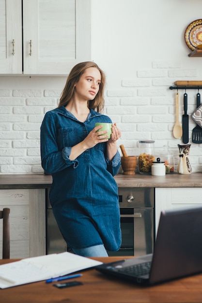 Junges hübsches schwangeres Mädchen arbeitet zu Hause im Selbstisolationsmodus in Quarantäne und macht eine Pause, um sich mit Kaffee auszuruhen