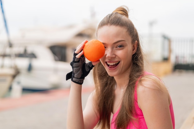 Junges hübsches Sportmädchen im Freien, das eine Orange mit überraschtem Gesichtsausdruck hält