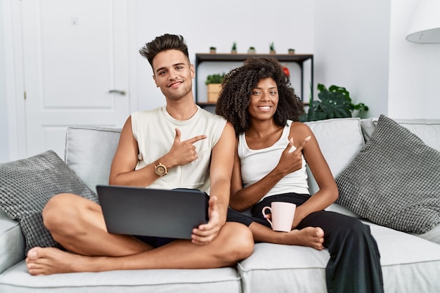 Junges interrassisches Paar mit Laptop zu Hause sitzend auf dem Sofa fröhlich mit einem Lächeln im Gesicht, das mit Hand und Finger zur Seite zeigt, mit glücklichem und natürlichem Ausdruck