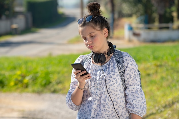 Junges Jugendlichmädchen, das den Schulbus wartet und ihren Smartphone verwendet