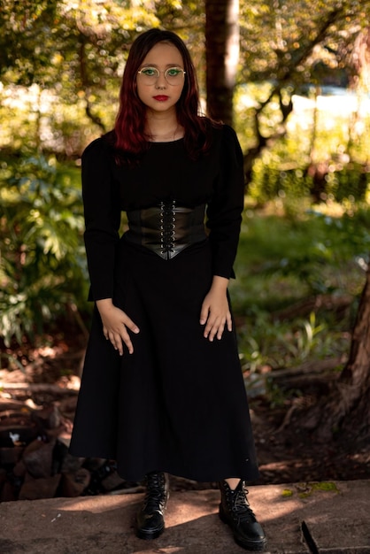 Junges kaukasisches dunkelhaariges Mädchen mit roten Streifen im schwarzen Kleideroutfit mit Korsettgürtel und Brille, das für weibliches Schönheitsshooting posiert