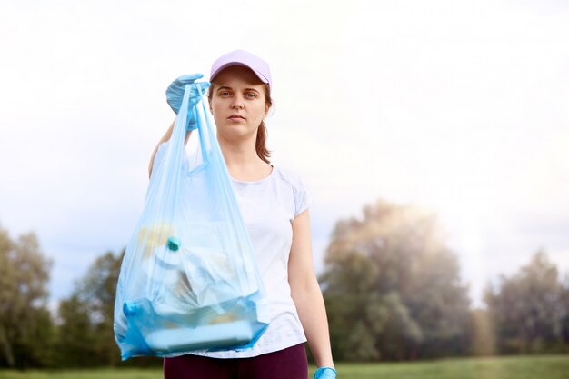 Junges kaukasisches Mädchen, das T-Shirt und Baseballmütze trägt, die draußen mit Müllsack aufwirft, kümmert sich um die Umwelt, stehend mit Himmel und Bäumen