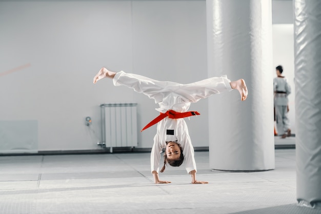 Junges kaukasisches Mädchen im Dobok, das Handstand mit gespreizten Beinen auf Taekwondo-Klasse tut.