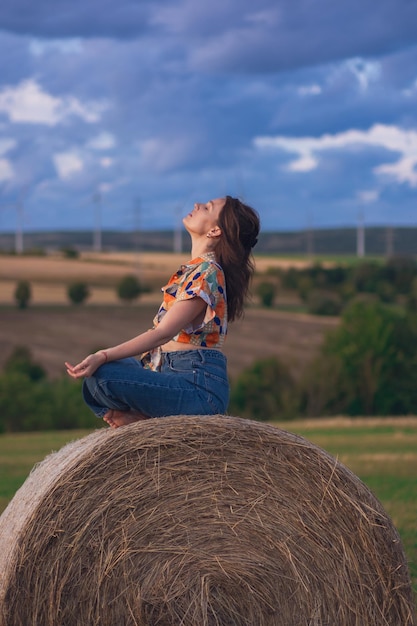 Junges kaukasisches Mädchen in Jeans und bunter Bluse sitzt auf einem runden Strohballen auf dem Feld. Langhaarige junge Frau posiert auf einem Feld vor bewölktem Himmel und Windmühlen