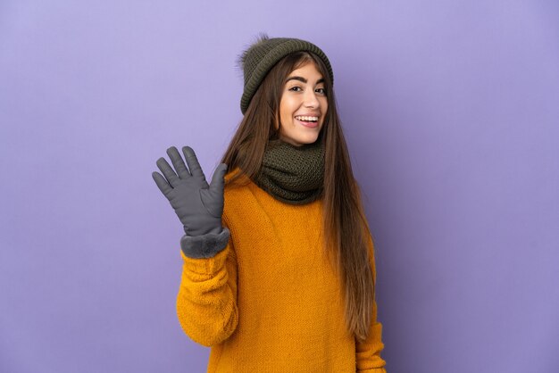 Junges kaukasisches Mädchen mit Wintermütze auf violettem Hintergrund, das mit der Hand mit glücklichem Ausdruck grüßt
