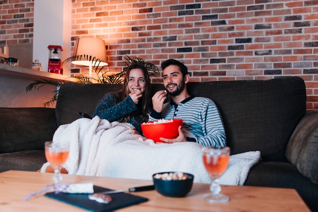 Junges kaukasisches Paar, das auf einem grauen Sofa sitzt, während der Fernseher Popcorn isst