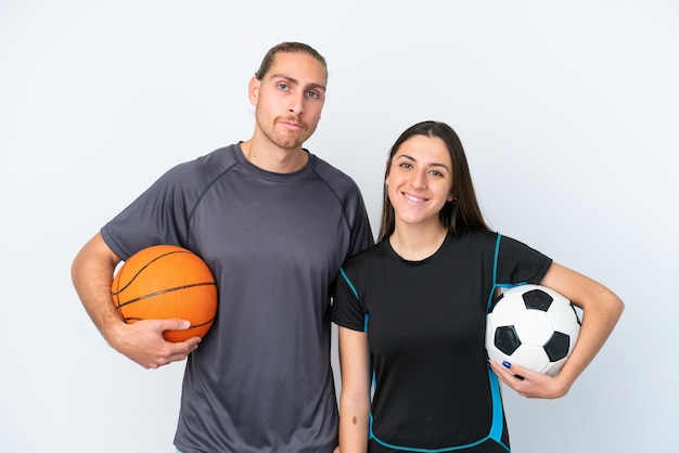 Junges kaukasisches Paar, das Basketball und Fußball spielt, isoliert auf weißem Hintergrund, posiert mit Armen an der Hüfte und lächelnd