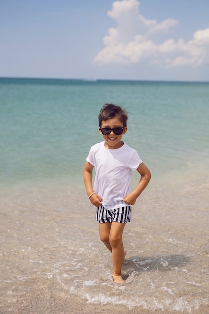Junges Kind in gestreiften Shorts und einem weißen T-Shirt geht am Sandstrand und in Sonnenbrille spazieren