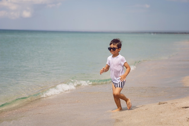 Junges Kind in gestreiften Shorts und einem weißen T-Shirt geht am Sandstrand und in Sonnenbrille spazieren
