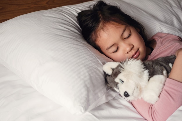 Junges kleines asiatisches Mädchen der gemischten Rasse, das im Bett mit ihrem Hundespielzeug, Schlafenszeitroutine, Weckkind für Schule, Kinderschlafstörung schläft