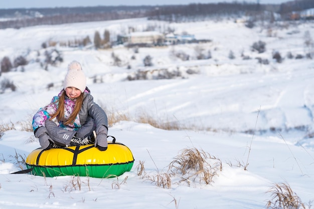 Junges kleines Mädchen, das im Winter auf dem schneebedeckten Hügel lacht und Snowtubing genießt