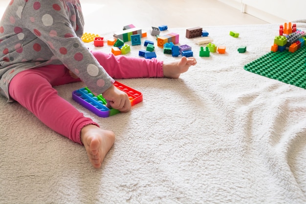 Junges kleines Mädchen, das zu Hause auf einem Teppich sitzt und mit neuem trendigem Spielzeug spielt, pop it