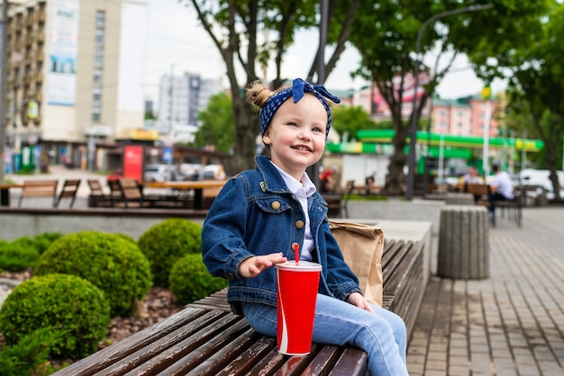 Junges kleines Mädchen mit Fast-Food-Beutel nahe Café, das auf der Bank sitzt