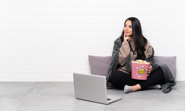 Junges kolumbianisches Mädchen, das eine Schüssel Popcorn hält und einen Film in einem Laptop denkt eine Idee beim oben schauen zeigt