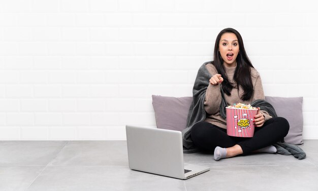 Junges kolumbianisches Mädchen, das eine Schüssel Popcorn hält und einen Film in einem Laptop überrascht zeigt und Front zeigt