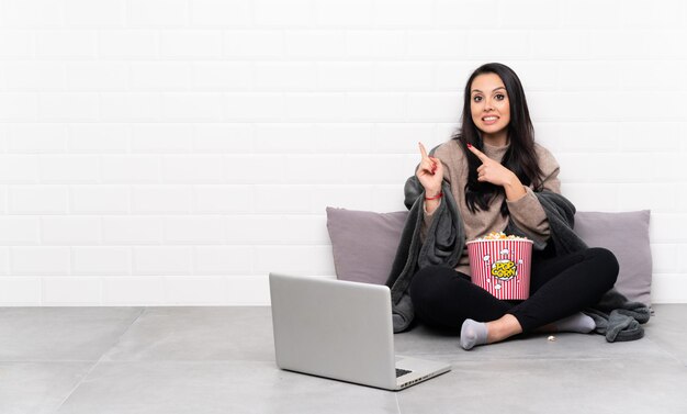 Junges kolumbianisches Mädchen, das eine Schüssel Popcorn hält und einen Film in einem Laptop zeigt, der erschrocken ist und zur Seite zeigt