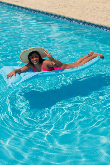 Junges lächelndes afroamerikanisches Mädchen im Bikini, mit Strohhut, der sich auf Schlauchboot im Schwimmbad entspannt.