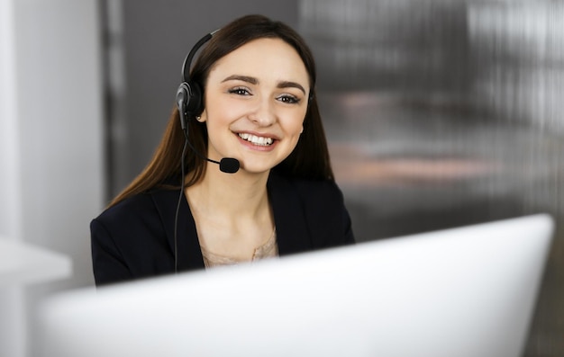 Junges lächelndes Mädchen in Headsets spricht mit dem Kunden der Firma, während sie am Schreibtisch im Büro eines Unternehmens sitzt. Porträt eines Call-Center-Betreibers bei der Arbeit.