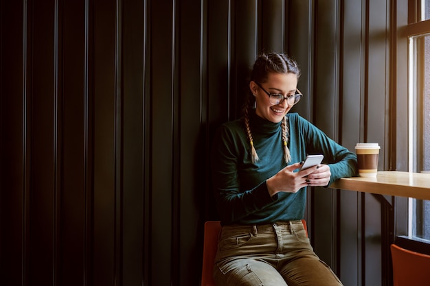 Junges lächelndes Mädchen mit Zöpfen, die im Kaffeehaus sitzen, eine Pause machen und Smartphone für das Aufhängen an sozialen Medien verwenden.