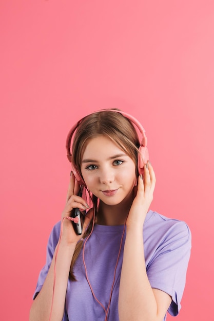 Junges lächelndes Mädchen mit zwei Zöpfen in lila T-Shirt, das Musik in Kopfhörern hört und glücklich in die Kamera blickt, über rosafarbenem Hintergrund isoliert
