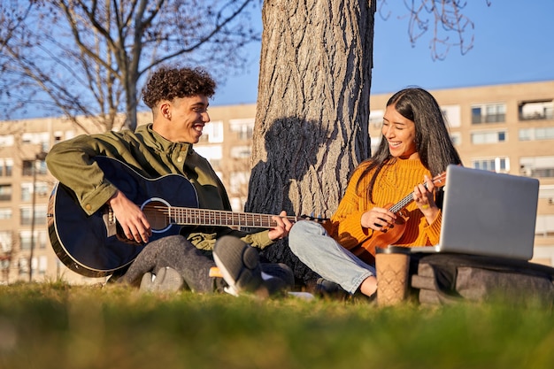 Junges lateinamerikanisches Paar, das am Baum sitzt und Ukulele und Gitarre spielt, singt ein Lied glückliches Gefühlentspannendes Konzept