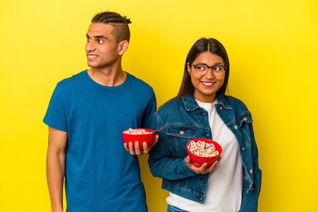 Junges lateinisches Paar, das eine Müslischale einzeln auf gelbem Hintergrund hält, sieht beiseite lächelnd, fröhlich und angenehm aus.