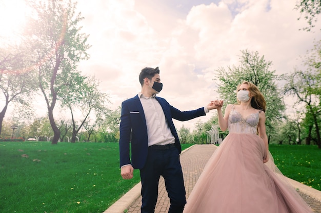 Junges Liebespaar, das während der Quarantäne an ihrem Hochzeitstag in medizinischen Masken im Park spazieren geht.