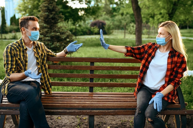 Junges Liebespaar in Masken und Handschuhen, die auf Bank im Park sitzen, Quarantäne. Romantisches Treffen während der Epidemie, Gesundheitsversorgung und Schutz, Pandemie Lebensstil