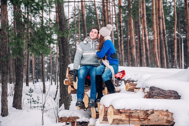 Junges Liebespaar kleidete in der blauen Strickjacke an, die auf dem Brennholz sitzt und im Winterwald umarmt