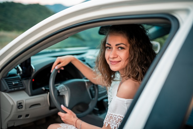 Junges lockiges reizendes Mädchen lächelt, ein weißes Auto im Freien fahrend