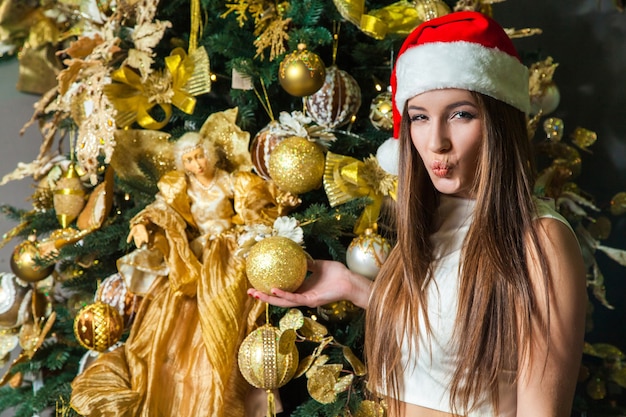 Junges lustiges schönes Modemodell mit dunklen Augen, braunem Haar und Weihnachtsmütze, das zu Hause das neue Jahr feiert. Dekoration des neuen Jahres, mit positiver Emotion, die auf die Kamera posiert und schaut. Gelbgold-Dekor..