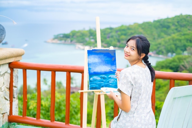 Junges Mädchen, das ein Pinselzeichnungsbild auf Leinwandpapier bei wunderschönem Landschaftsblick auf Koh Tao Thailand verwendet