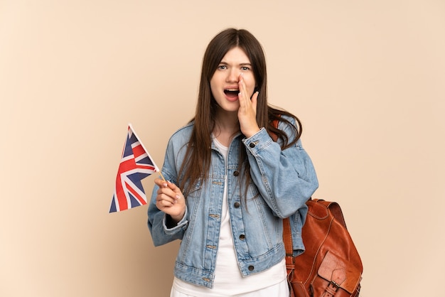 Junges Mädchen, das eine britische Flagge lokalisiert auf Beige mit Überraschung und schockiertem Gesichtsausdruck hält