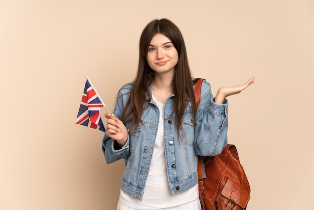 Junges Mädchen, das eine Flagge des Vereinigten Königreichs lokalisiert auf Beige hält, die Zweifel beim Erhöhen der Hände hat