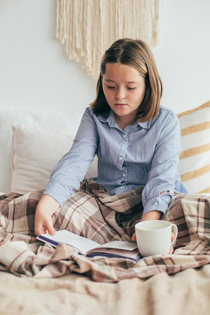 Junges mädchen, das eine gemütliche tasse mit kakao oder schokolade hält. junge frau im grauen strickpullover, der auf dem bett sitzt. gemütliches zuhause, hygge, komfort, stimmungskonzept