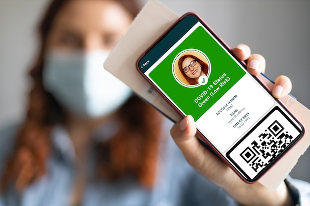 Junges Mädchen, das eine Gesichtsmaske trägt, die einen Reisepass, einen Ticketpass und ein Smartphone mit einer digitalen Gesundheitspass-App für Reisen während der Covid-19-Pandemie hält.