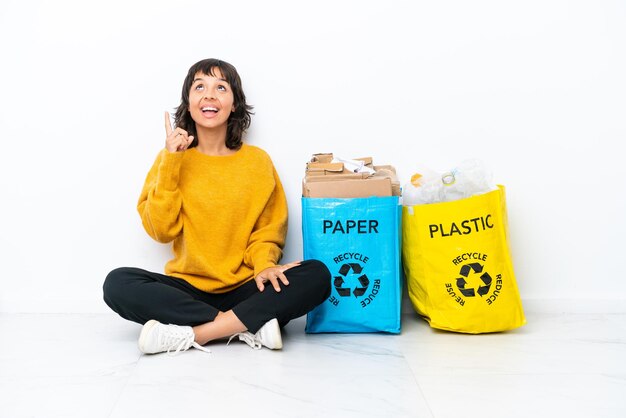 Junges Mädchen, das eine Tasche voller Plastik und Papier hält, die auf dem Boden sitzt, isoliert auf weißem Hintergrund, nach oben zeigend und überrascht