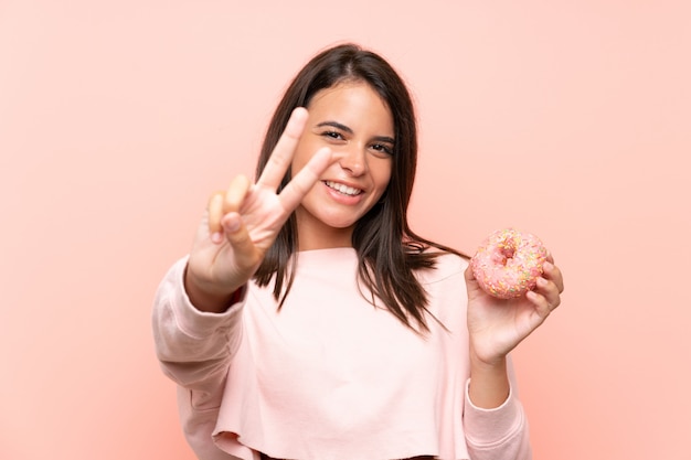 Junges Mädchen, das einen Donut über lokalisierter rosa Wand lächelt und zeigt Siegeszeichen hält