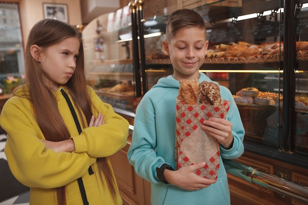 Junges Mädchen, das ihren Bruder wütend ansieht, während er frisch gebackenes Brot genießt