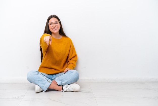 Junges Mädchen, das isoliert auf weißem Hintergrund auf dem Boden sitzt, zeigt mit einem selbstbewussten Ausdruck mit dem Finger auf dich