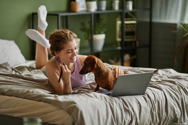 Junges Mädchen, das Laptop benutzt und mit ihrem Hund spielt, während es sich in der Freizeit zu Hause im Bett entspannt