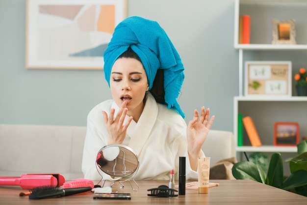 junges Mädchen, das Lipgloss umwickelte Haare in Handtuch anwendet, das am Tisch mit Make-up-Tools im Wohnzimmer sitzt?