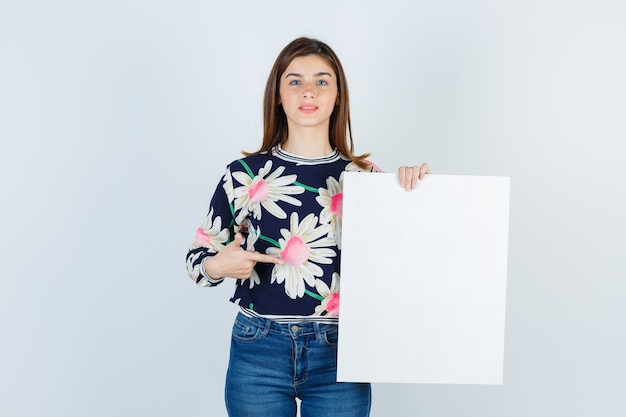 Junges Mädchen, das Papierplakat in der Blumenbluse, in den Jeans zeigt und verwirrt, Vorderansicht schaut.