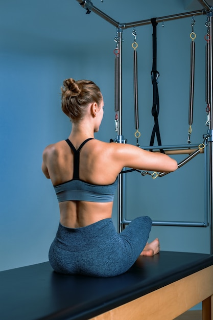 Junges Mädchen, das Pilates-Übungen mit einem Reformerbett macht. Schöner schlanker Fitnesstrainer auf grauer Wand des Reformators, zurückhaltend, Kunstlicht. Fitness-Konzept.