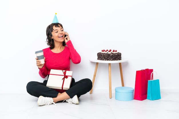 Junges Mädchen, das seinen Geburtstag feiert, das auf dem Boden sitzt, isoliert auf weißem Hintergrund, der Kaffee zum Mitnehmen und ein Handy hält