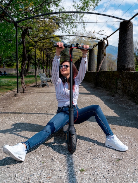 Junges Mädchen, das Spaß hat, ein Hemd und Kopfhörer zu tragen und einen E-Scooter in einem Park zu fahren?