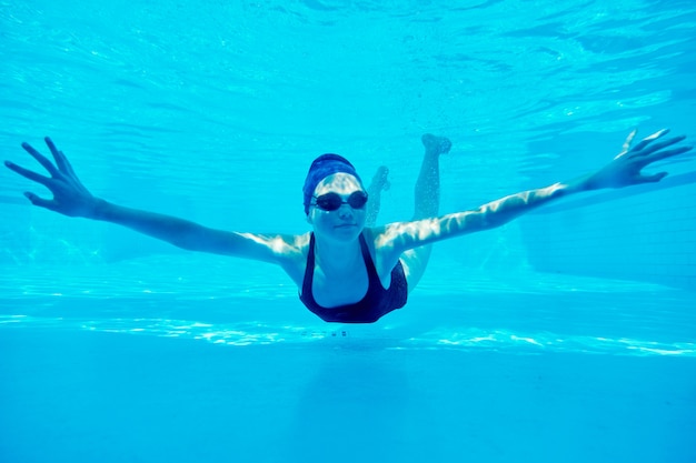 Junges Mädchen, das unter Wasser im Pool schwimmt, weiblich schaut und lächelt