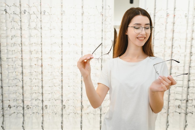 Junges Mädchen, das vor dem Spiegel eine Brille anprobiert Hilft, das Sehvermögen zu verbessern und die Gesundheit der Augen zu retten In der Nähe eines transparenten Ständers mit optischen Linsen stehen