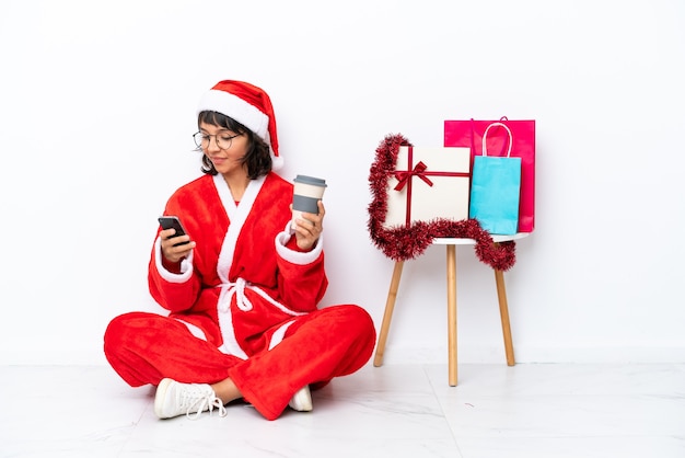 Junges Mädchen, das Weihnachten feiert, das auf dem Boden sitzt, isoliert auf weißem Hintergrund, der Kaffee zum Mitnehmen und ein Handy hält