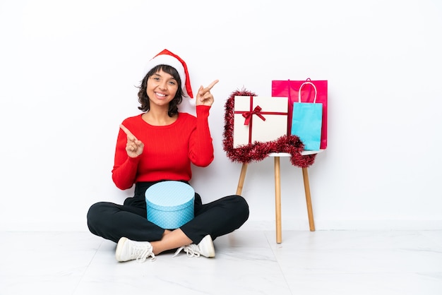 Junges Mädchen, das Weihnachten feiert, das auf dem Boden sitzt, isoliert auf weißem Hintergrund, der mit dem Finger auf die Seitenwände zeigt und glücklich ist